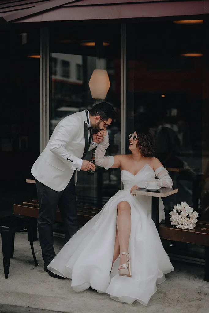 A groom in a white tuxedo jacket and black pants leaning in to kiss a bride sitting on a bench outside a building, wearing a white wedding dress, sunglasses, and high heels, with a bouquet of white flowers nearby.