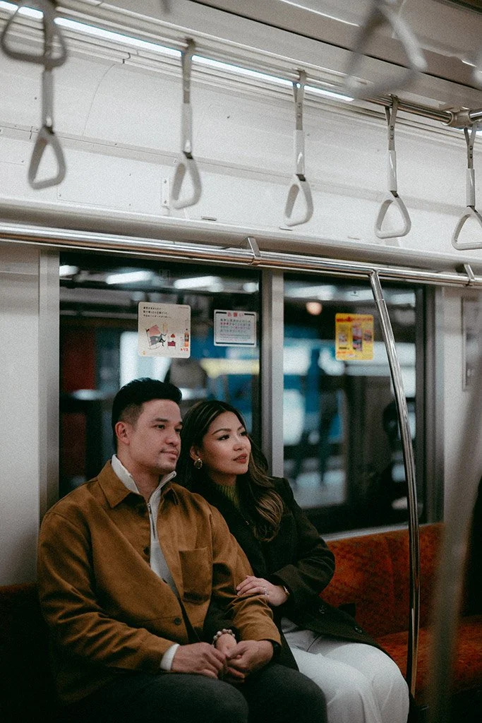 A man and woman sitting together in a subway train, holding hands and looking at something outside the frame. The man has short dark hair and is wearing a brown jacket, while the woman has long dark hair and is wearing a black coat.