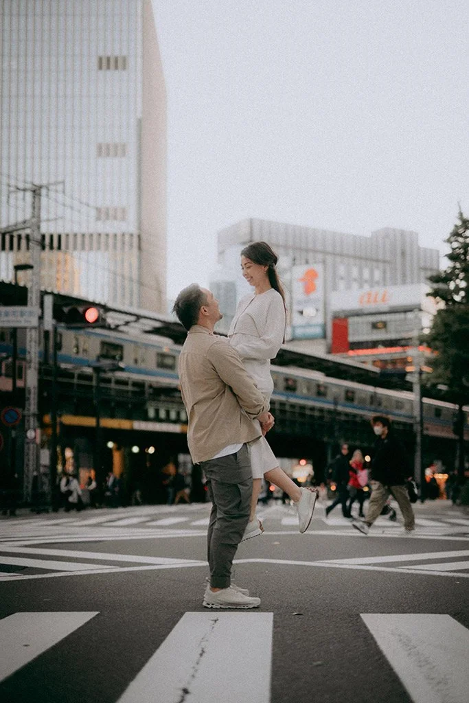 A man holding a woman in his arms in the middle of a busy city crosswalk during daytime, with buildings, pedestrians, and a train in the background.