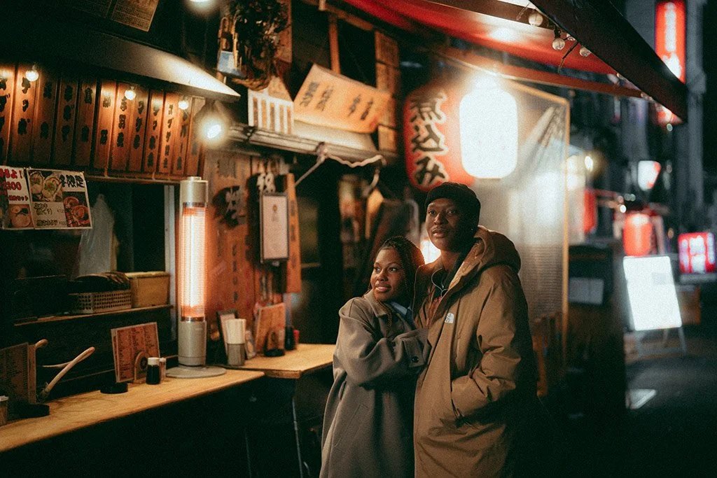 A couple stands close together outside a Japanese restaurant, illuminated by warm lighting and traditional lanterns, at night.