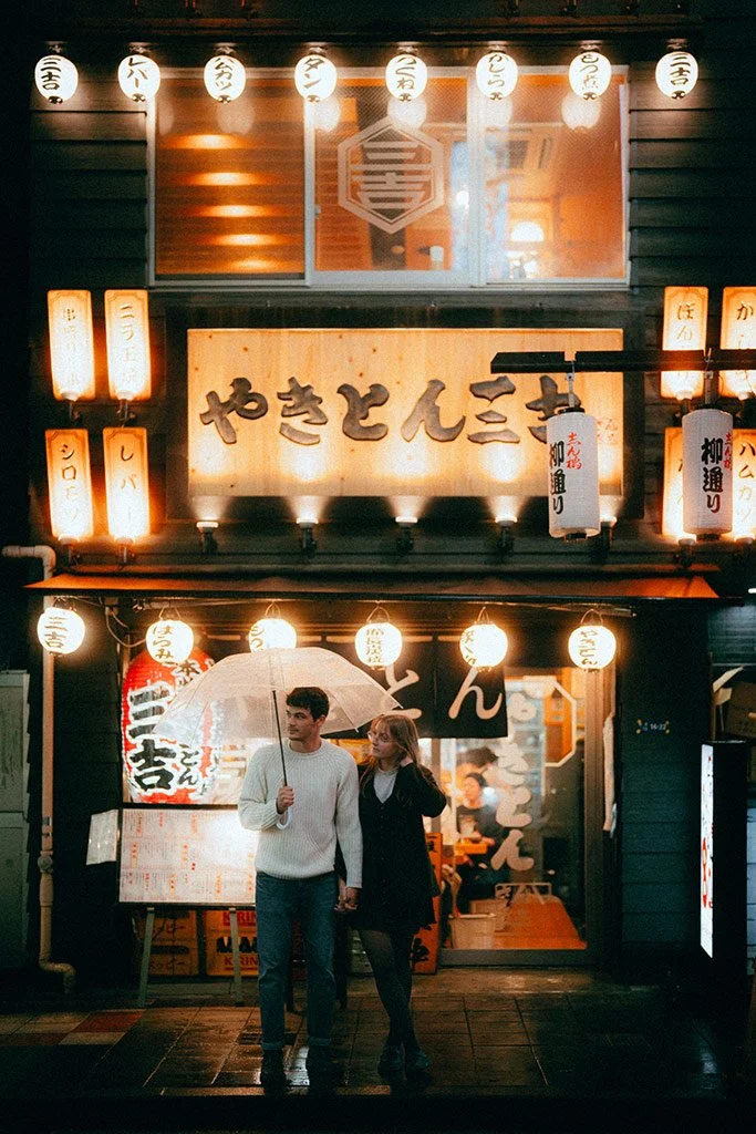 A couple walks together under an umbrella in front of a Japanese restaurant entrance decorated with paper lanterns. The restaurant signage has Japanese writing, with warm lighting illuminating the exterior at night.