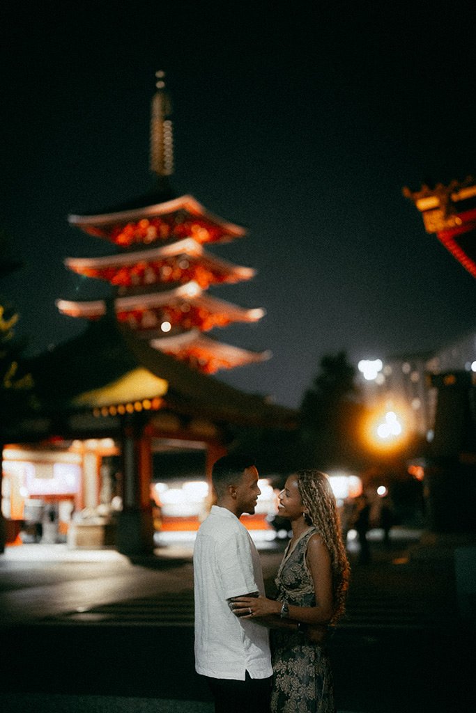 A couple stands close at night in front of a Japanese pagoda with red roofs, illuminated by warm lights, and city lights in the background.