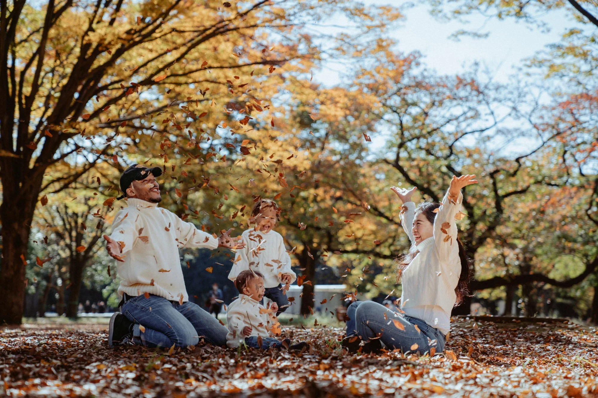 A group of people, including children and adults, playing and throwing autumn leaves in a park with trees that have colorful fall foliage.