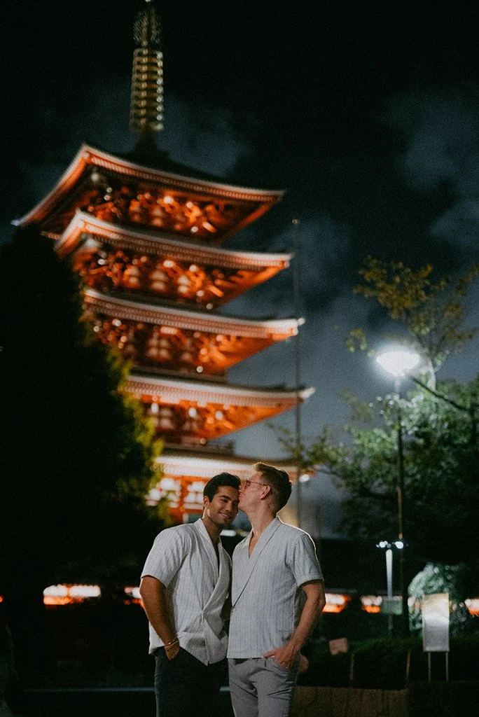 Two men standing close together at night in front of a traditional Japanese pagoda, with one man kissing the other's cheek.