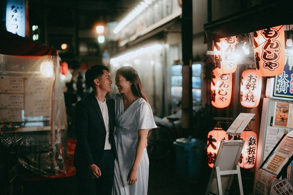 A couple in formal attire at a Japanese street food stall at night with illuminated lanterns and signage.