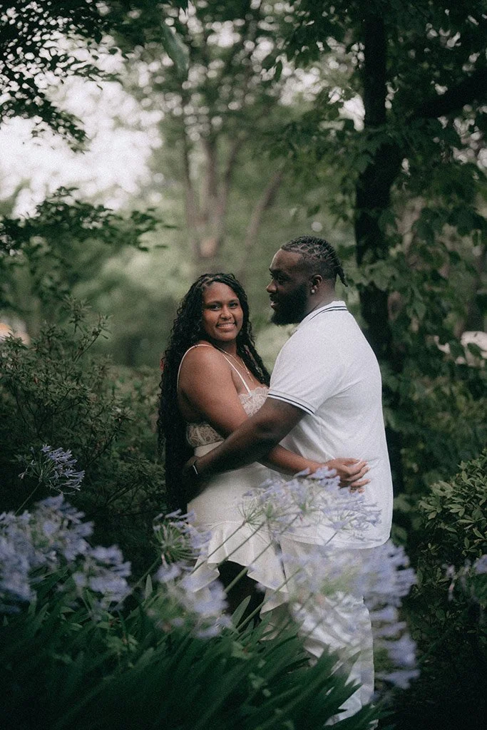 A couple standing close together outdoors among lush green trees and purple flowers, smiling and embracing.