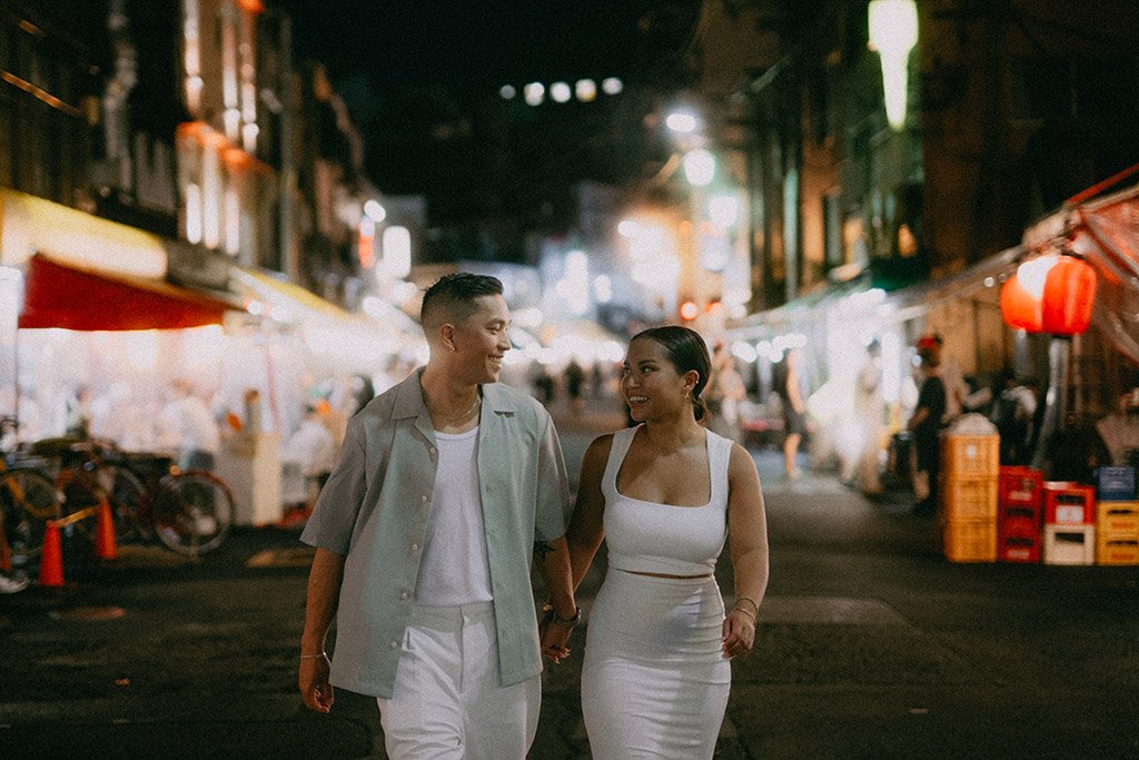 A young couple walking hand in hand through a lively, brightly lit street at night, with market stalls and other people in the background.