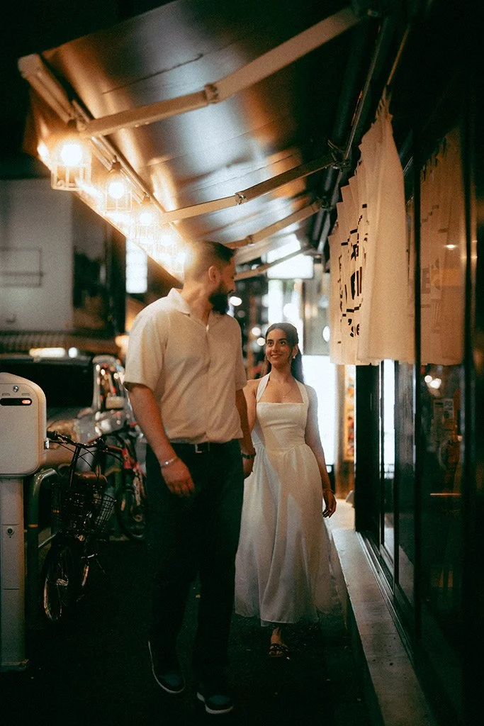 A man and woman walk together on a city sidewalk at night, illuminated by warm string lights, with bicycles and storefronts visible in the background, creating a cozy urban scene.
