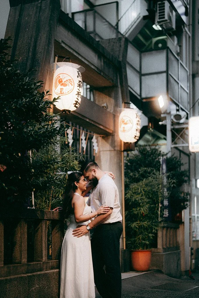 A couple stands close together on a city street at night, embracing, with Asian lanterns hanging above, and greenery in pots behind them.