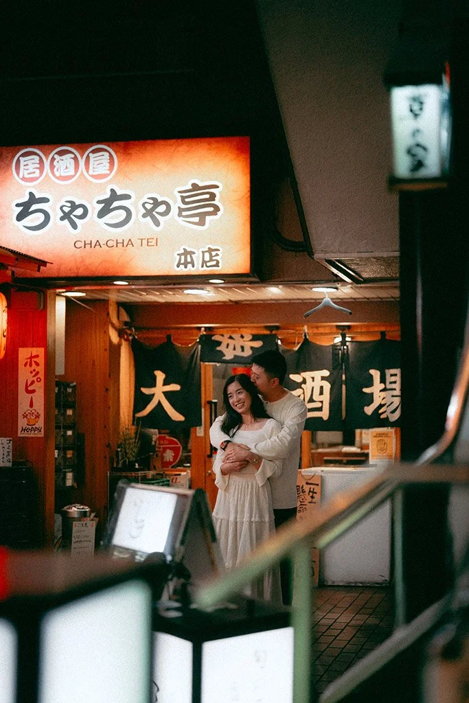 A smiling woman in a white dress embraces a man from behind inside a Japanese restaurant with hanging black noren curtains and illuminated signs in Japanese