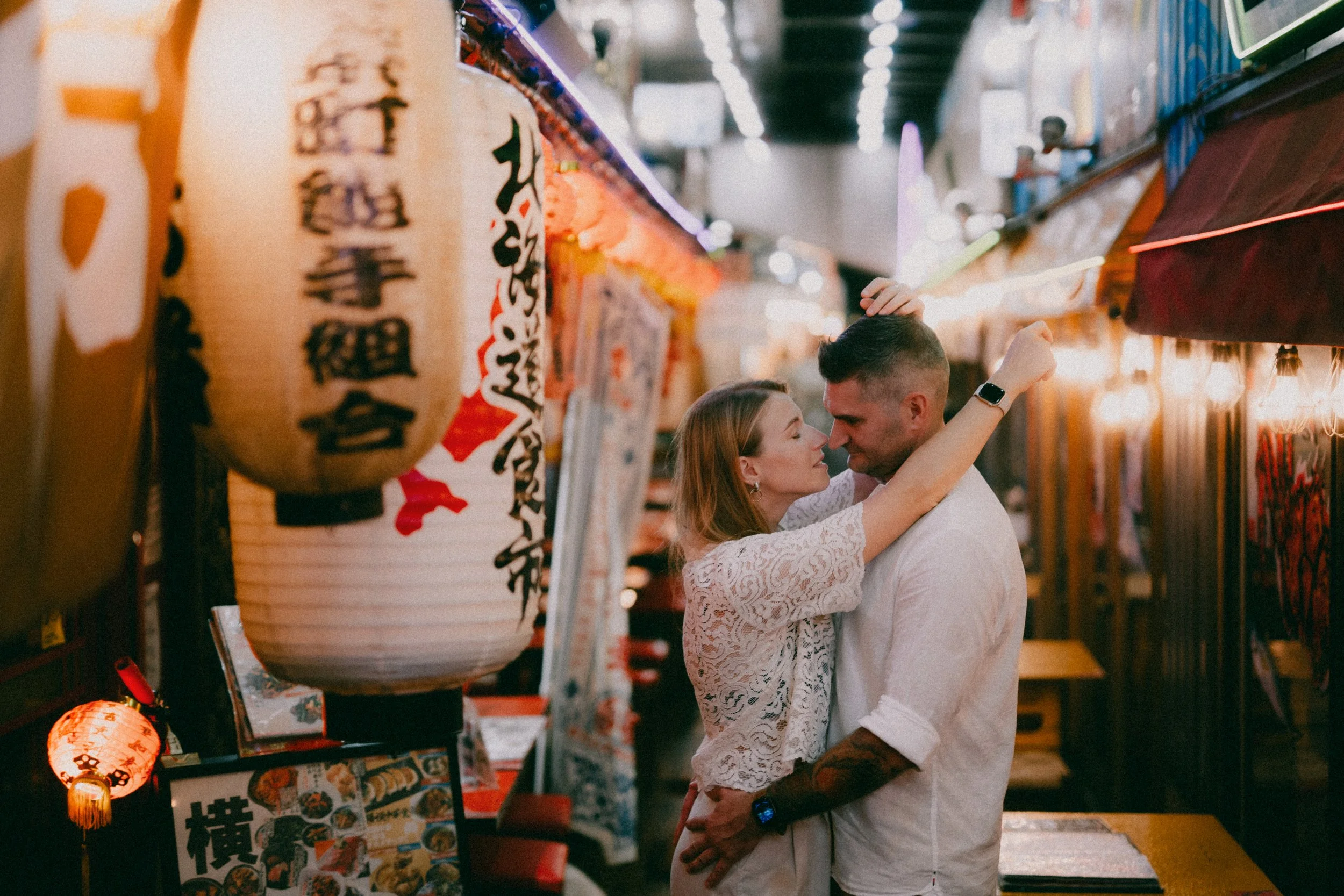 A couple is dancing closely inside a Japanese-themed market or shop. The woman is wearing a white lace dress, and the man is dressed in a white shirt with rolled-up sleeves and has tattoos on his arms. They are smiling and looking into each other's e