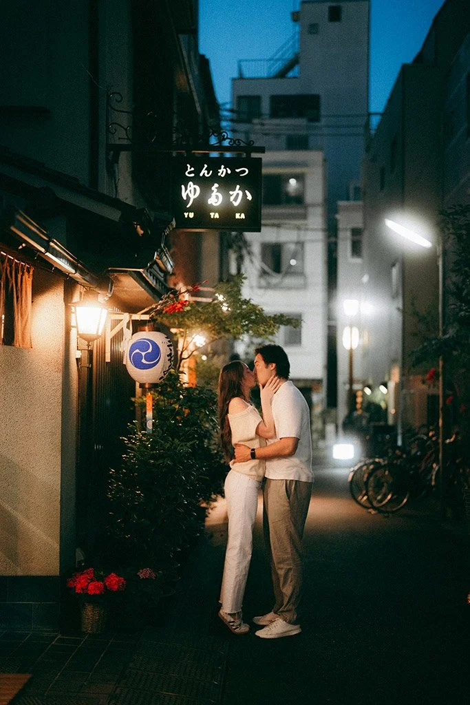 A couple kissing on a dimly lit street at night, with buildings and bicycles in the background, Japanese signs and paper lanterns are visible.