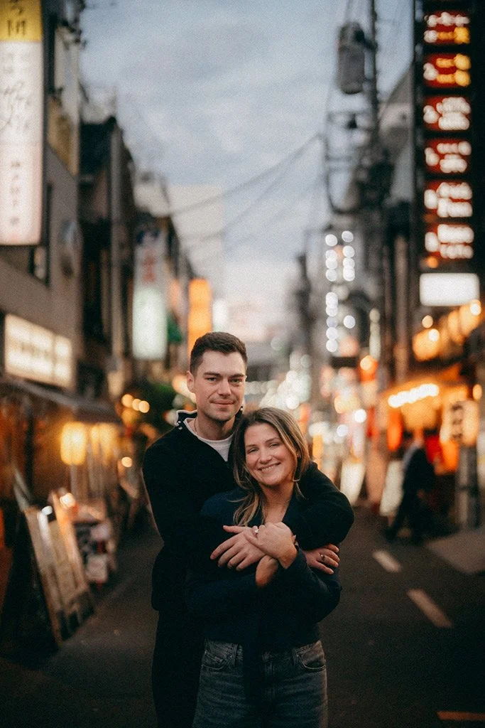 A smiling young couple hugging on a lively urban street at dusk with neon signs and shops in the background.