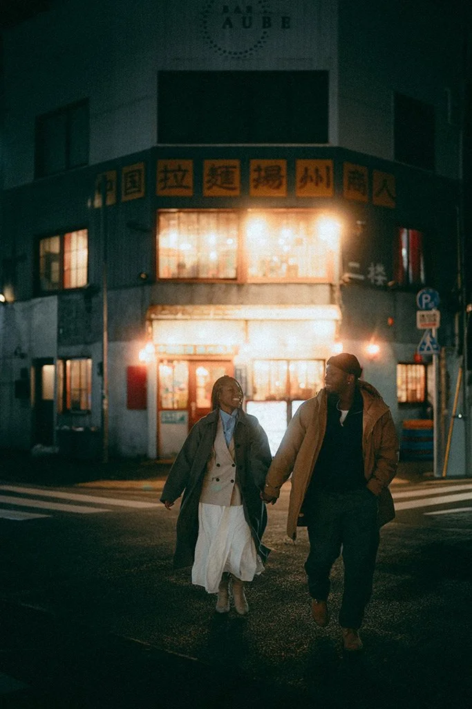 A couple holding hands and walking at night on a city street, illuminated by warm streetlights, in front of a building with bright signs.