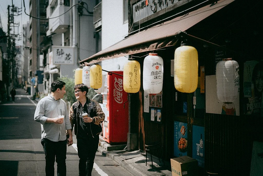 Two men walking and talking on a narrow street in Japan, with paper lanterns hanging outside a small shop or restaurant.