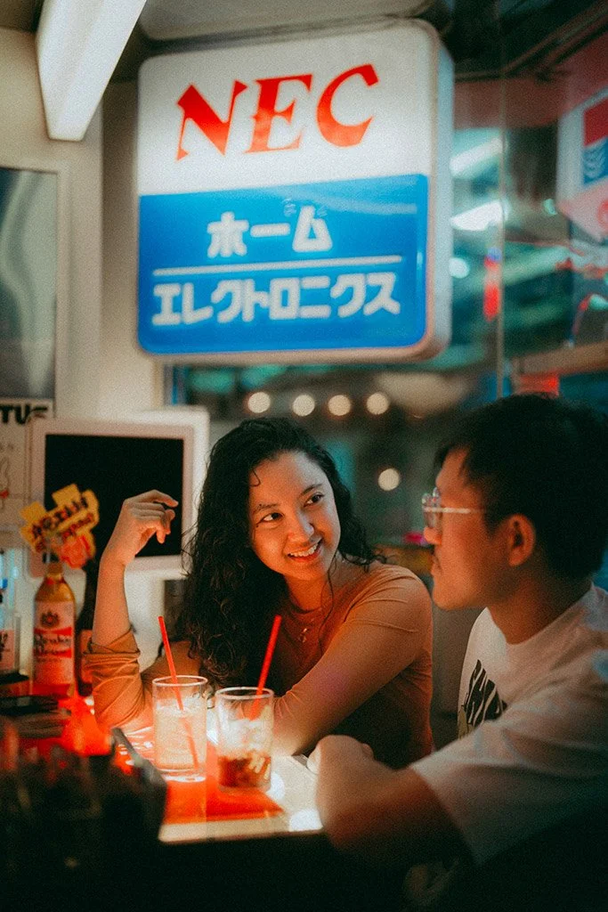 Two young adults, a woman with dark curly hair and a man with glasses, are sitting at a table in a bar or restaurant, smiling and talking. There are drinks with red straws on the table. The background displays neon signs with Japanese text, indicatin