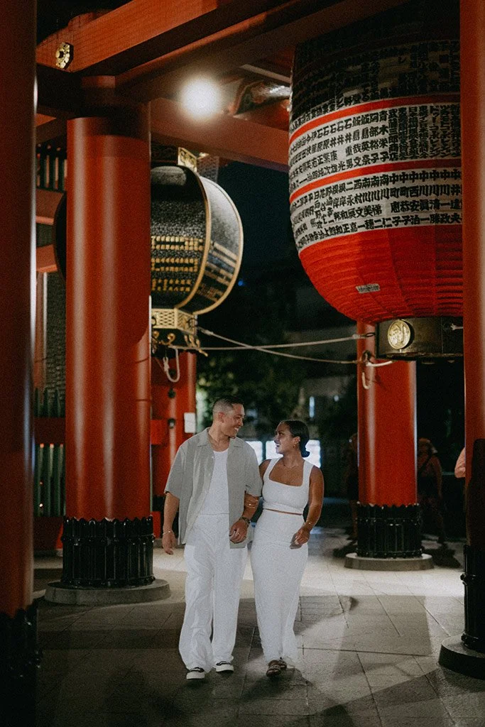 A couple dressed in white walking together at night under traditional Japanese lanterns and large drum decorations.
