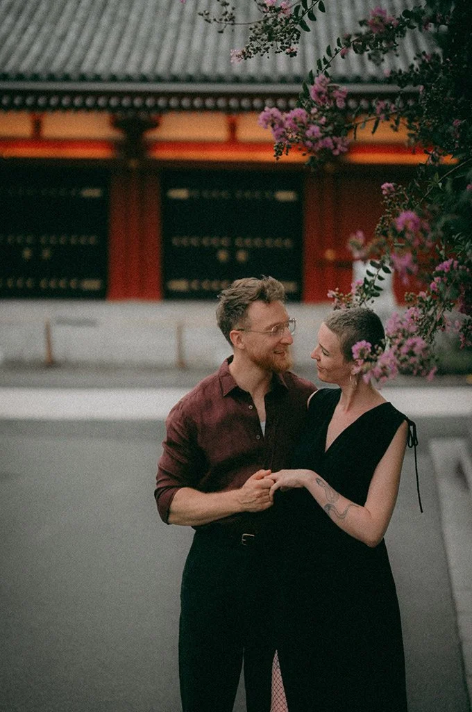 A couple holding hands and gazing at each other outdoors, with a traditional Asian-style building and pink flowering branches in the background.