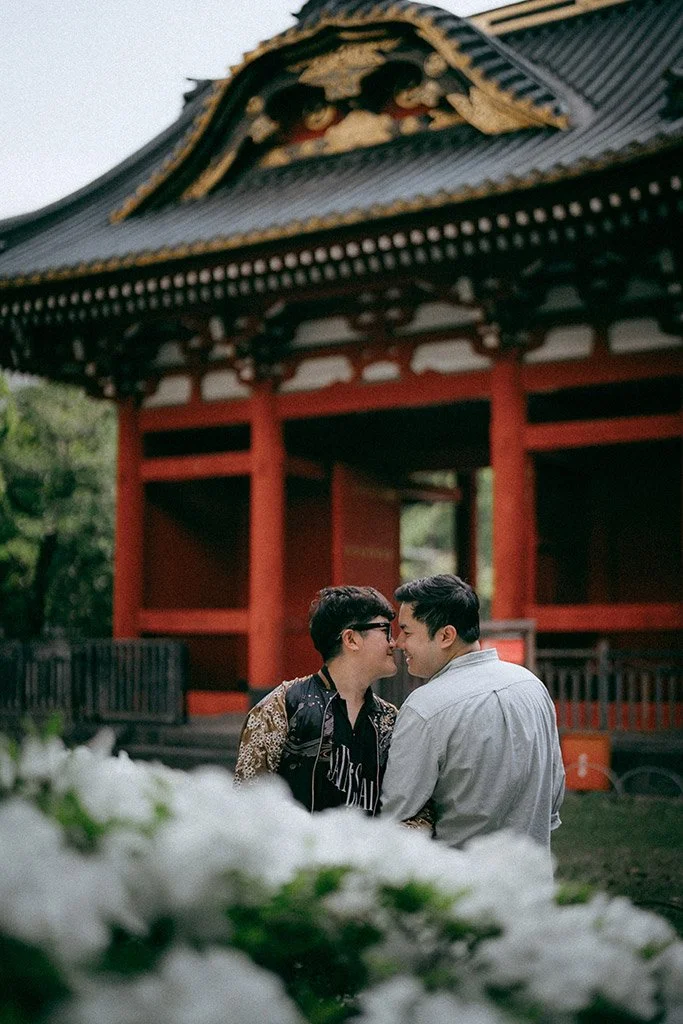 Two men sharing a tender moment, close to each other with foreheads touching, in front of a traditional Asian pavilion with red columns and a decorative roof.