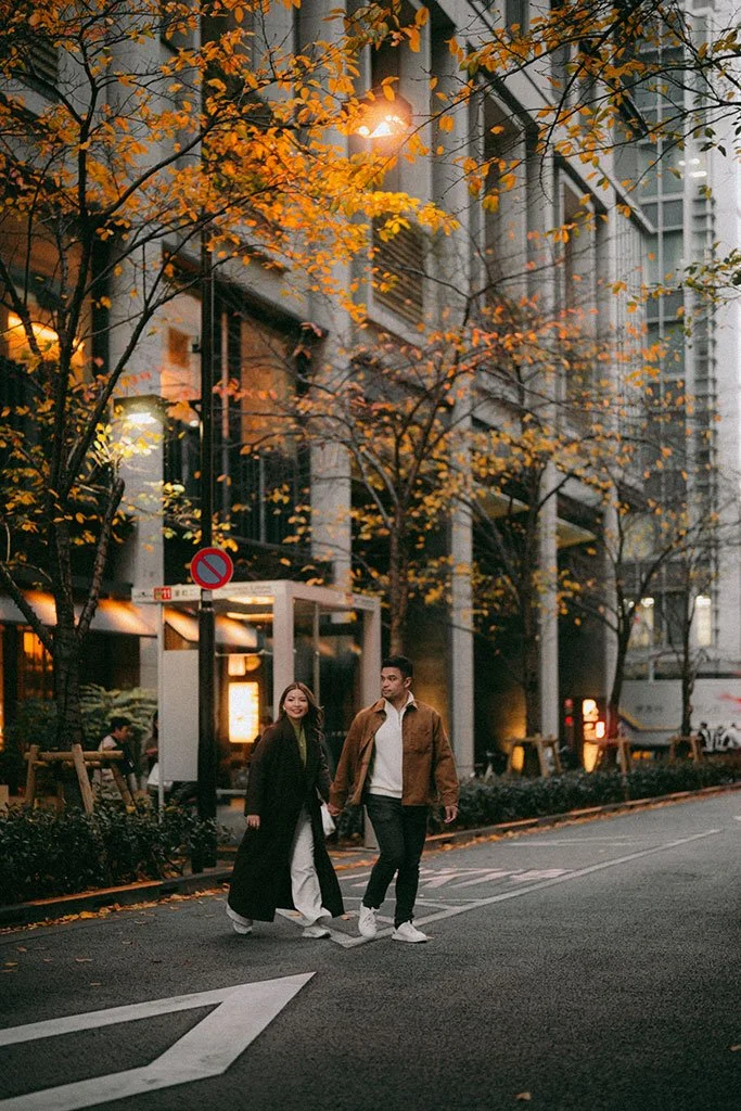 A young couple walking hand-in-hand on a city street during autumn evening, with trees with orange leaves and a modern building in the background.