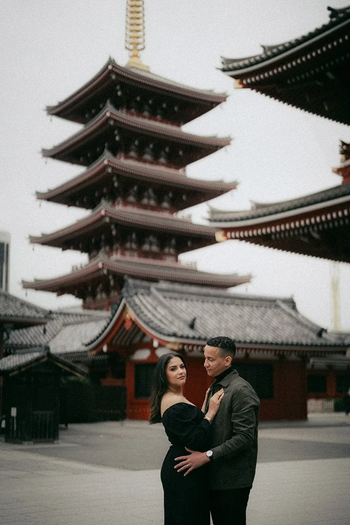 A couple stands together in front of a traditional Japanese pagoda, with the woman in a black dress and the man in a dark jacket, during daylight hours.