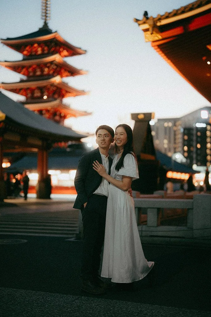 A young couple stands together outside at dusk, with traditional Japanese pagodas and buildings illuminated in the background.