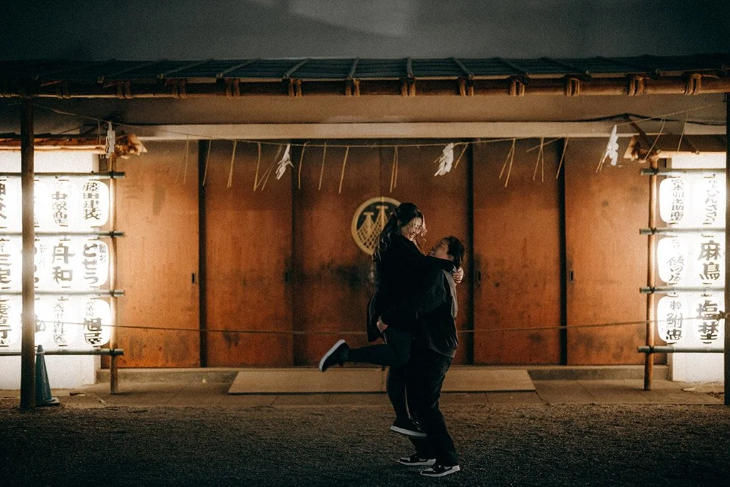 Two people embrace and smile in front of a traditional Japanese building illuminated with lanterns at night.