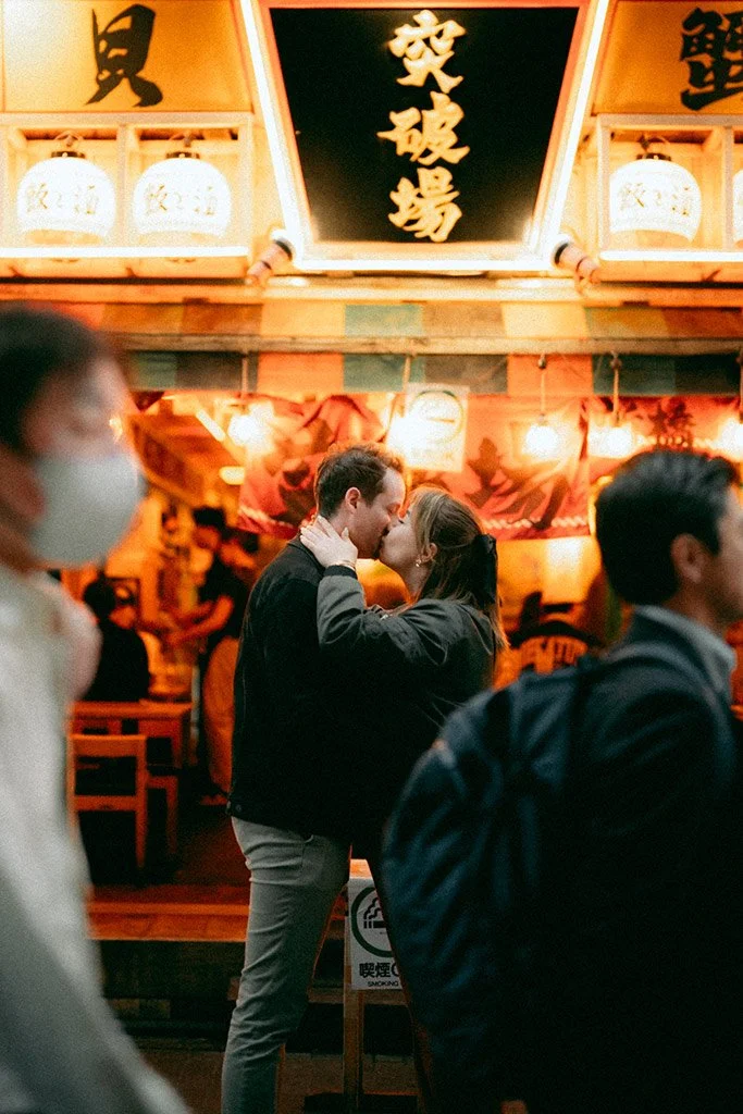 A couple kissing in a crowded, warmly lit outdoor restaurant or shopping area with Japanese lanterns and traditional decor.