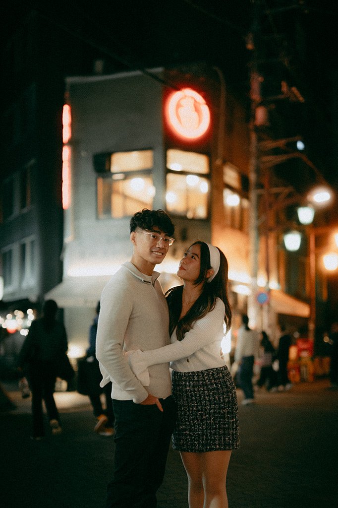 A young couple standing close together on a city street at night, with city lights and a building with illuminated signage in the background.