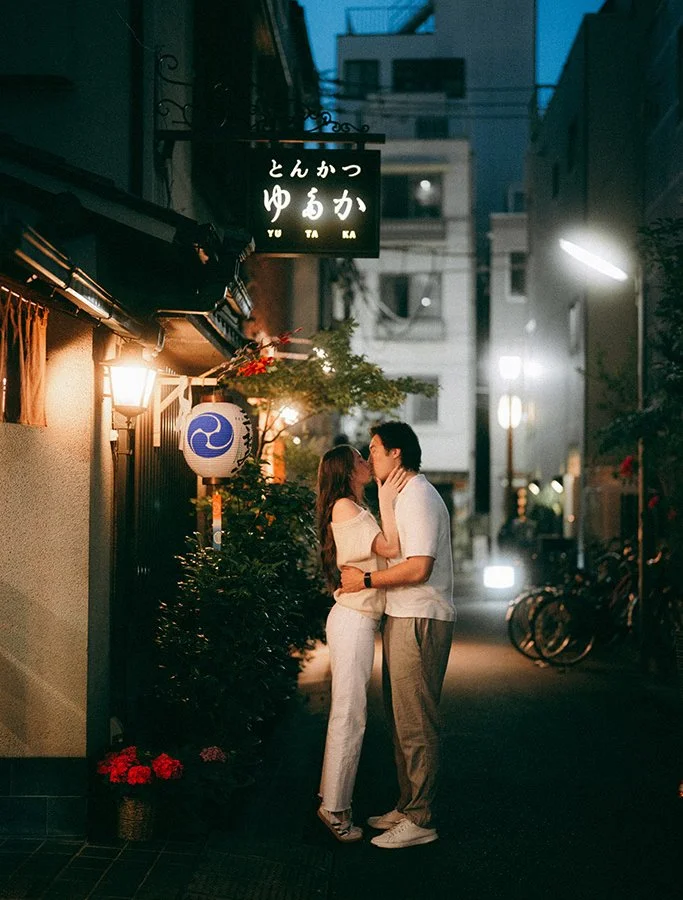 A couple kissing on a narrow street at night, illuminated by streetlights, with a Japanese restaurant sign overhead, bicycles parked along the side, and buildings in the background.