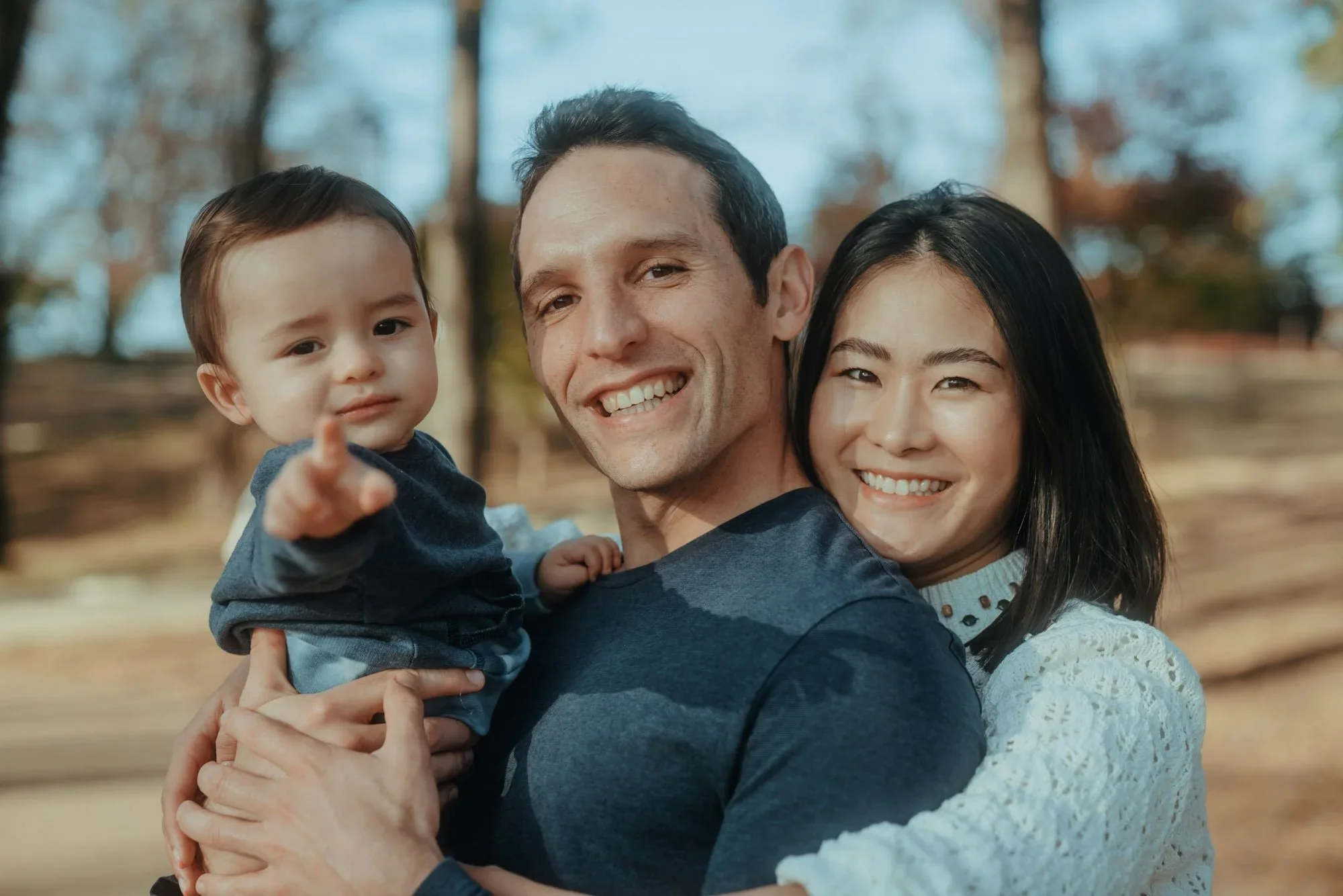 A happy family of three outdoors in a park during autumn, with trees in the background. The father is holding their young son, and the mother is standing beside them. All are smiling and looking at the camera.