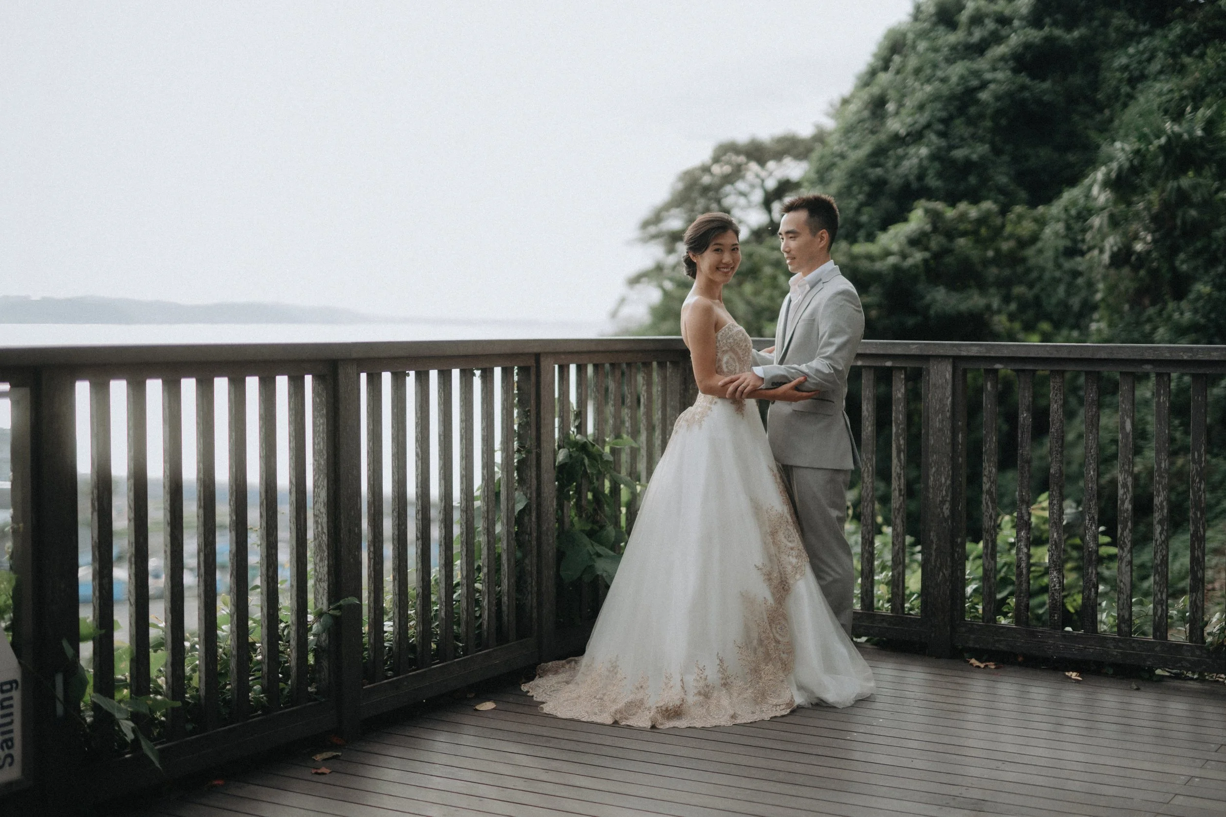 A bride and groom dressed in wedding attire standing on a wooden deck with a railing, holding hands and smiling at each other. The bride wears a strapless white wedding gown, and the groom wears a light gray suit with a white shirt. There are trees a
