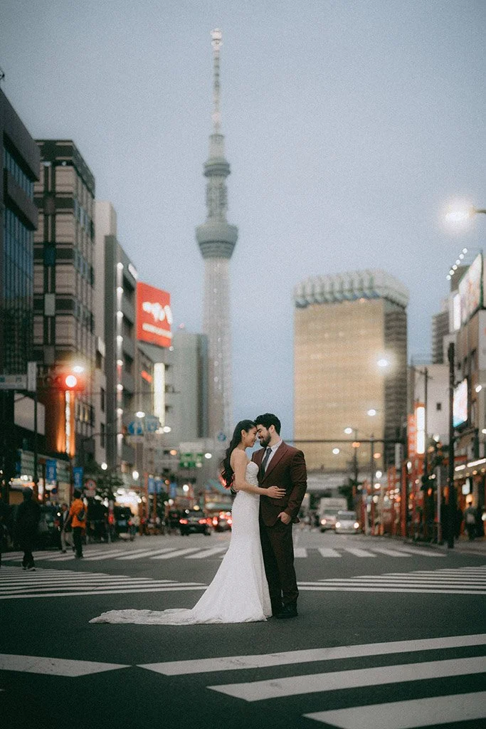 A bride and groom embrace on a city crosswalk at dusk, with the Tokyo Skytree tower in the background.