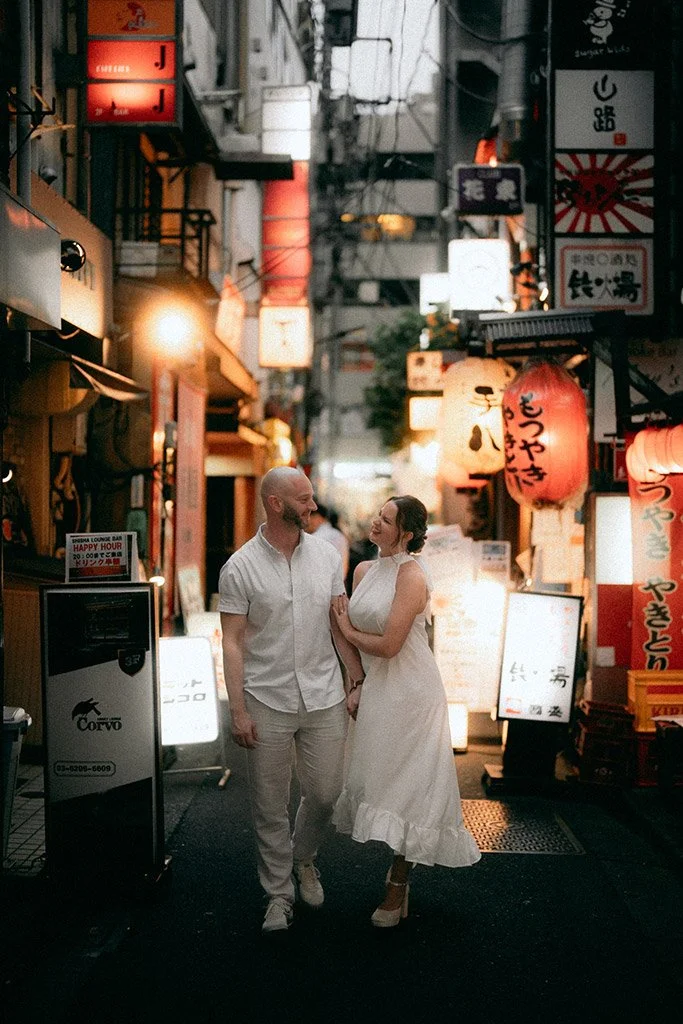 A couple holding hands and smiling at each other while walking through a narrow illuminated street in Japan, decorated with Japanese lanterns and signs.