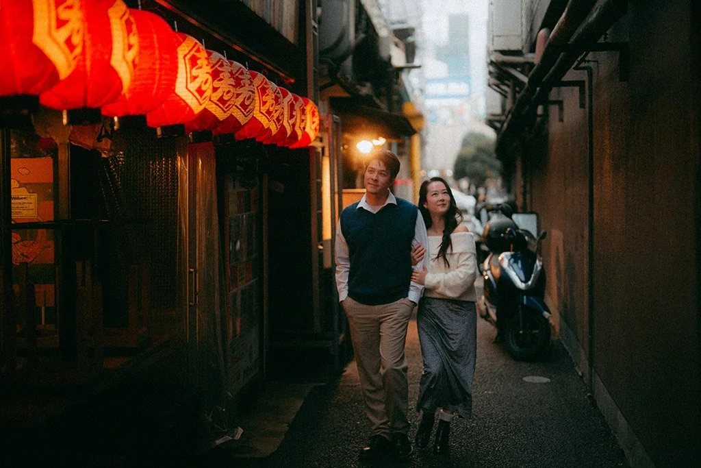 A couple walking arm in arm on a narrow street at dusk, with red lanterns hanging on the left side and buildings on both sides, one with motorcycle parked outside.