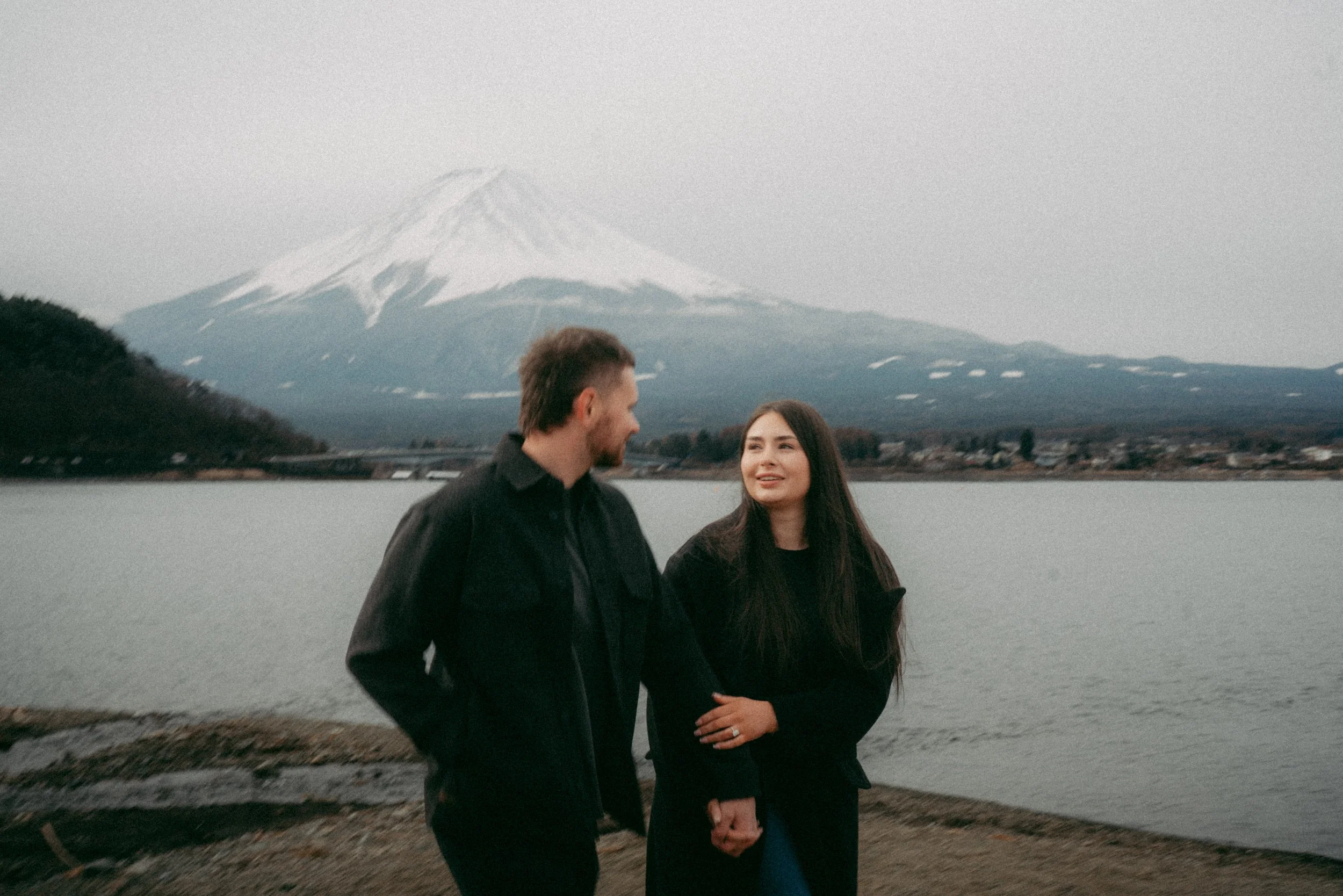 A man and woman standing by a body of water with a snow-capped mountain in the background, engaging in conversation.