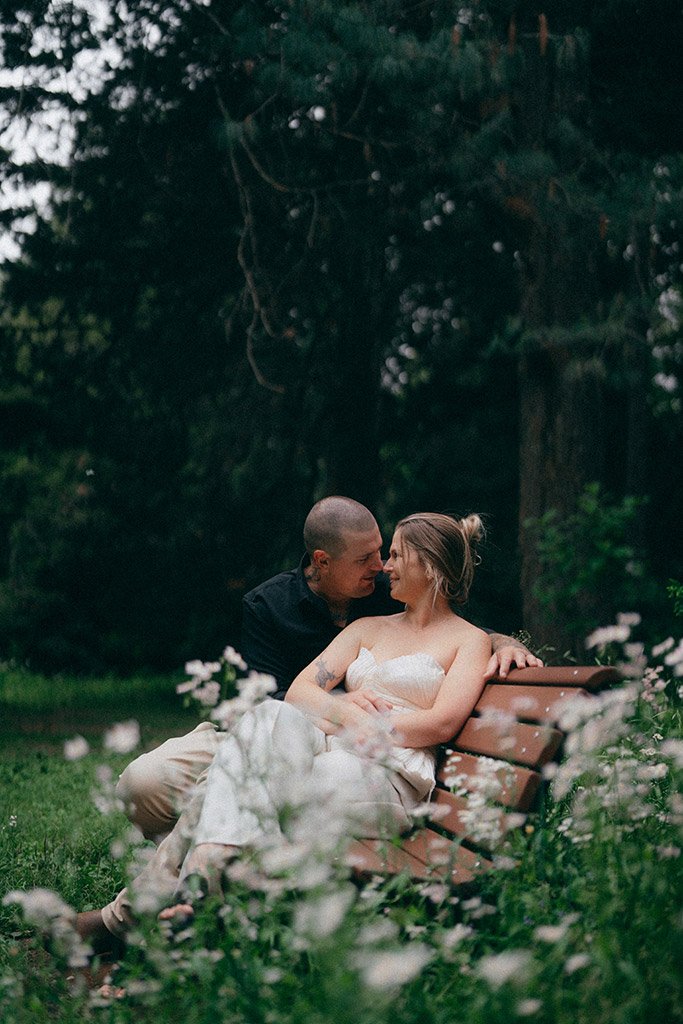 A couple sitting closely on a park bench surrounded by flowers, with trees in the background, during what appears to be evening.