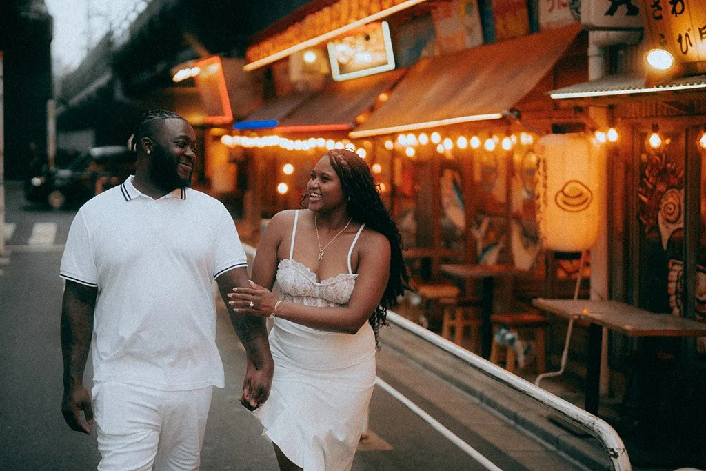 A smiling couple walking hand-in-hand on a city street at dusk, with illuminated storefronts and colorful signs in the background.