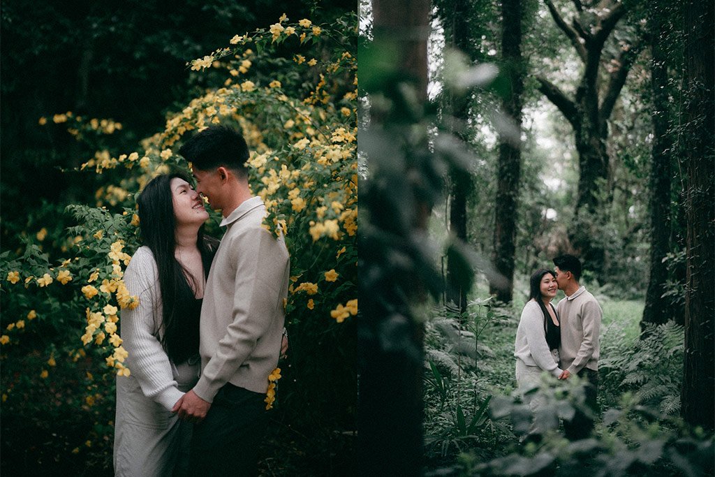 A couple standing close together in a lush green forest, smiling and holding hands. The woman has long black hair and is wearing a white sweater and dark pants, while the man has short dark hair and is wearing a light gray jacket and dark pants. They