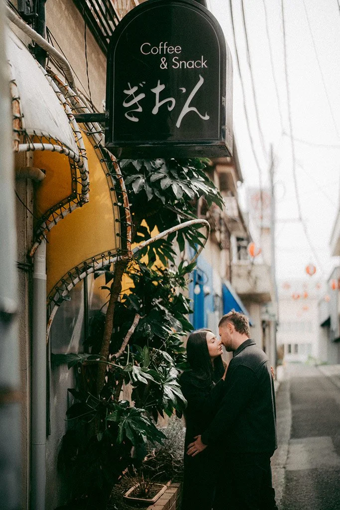 A couple stands close together on a narrow street, about to kiss, near a sign that says "Coffee & Snack" and Japanese characters, with plants and buildings in the background.