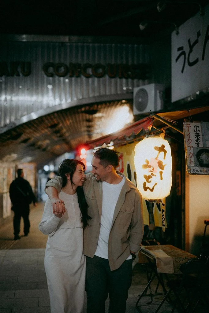 A happy couple walking arm-in-arm at a nighttime outdoor market or street in Japan, illuminated by traditional lanterns and signs with Japanese writing.
