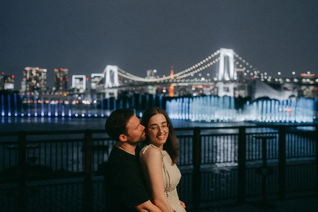 A couple smiling and embracing at night with a brightly lit suspension bridge and city skyline in the background.