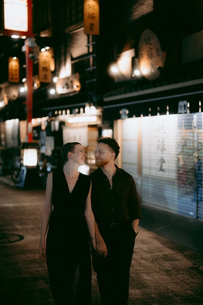 A couple walking hand in hand at night in a street with illuminated signs and lanterns, smiling and enjoying each other's company.