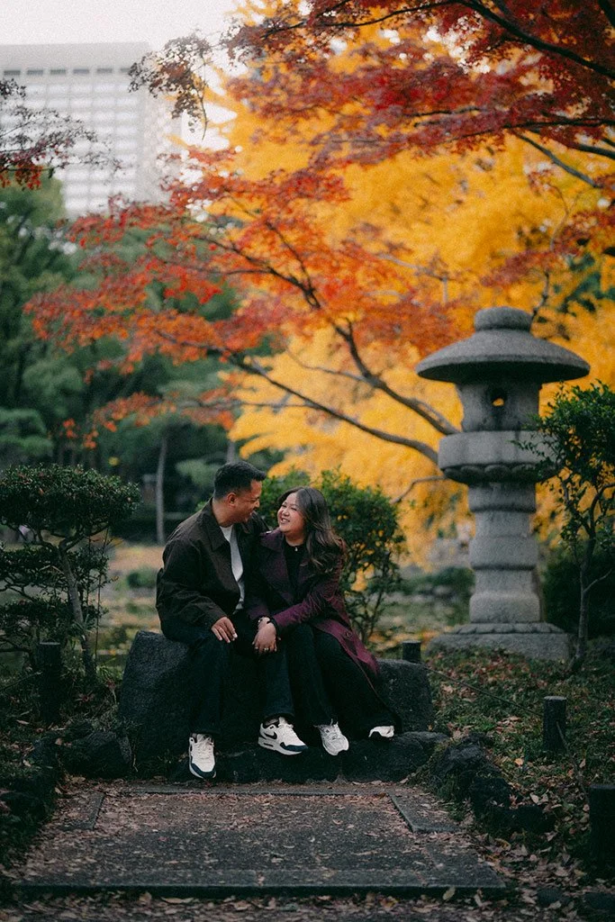 A couple sitting on a rock bench in a park during autumn, smiling and laughing together. There are vibrant fall foliage trees with orange, yellow, and red leaves, a stone lantern, and some bushes surrounding them.