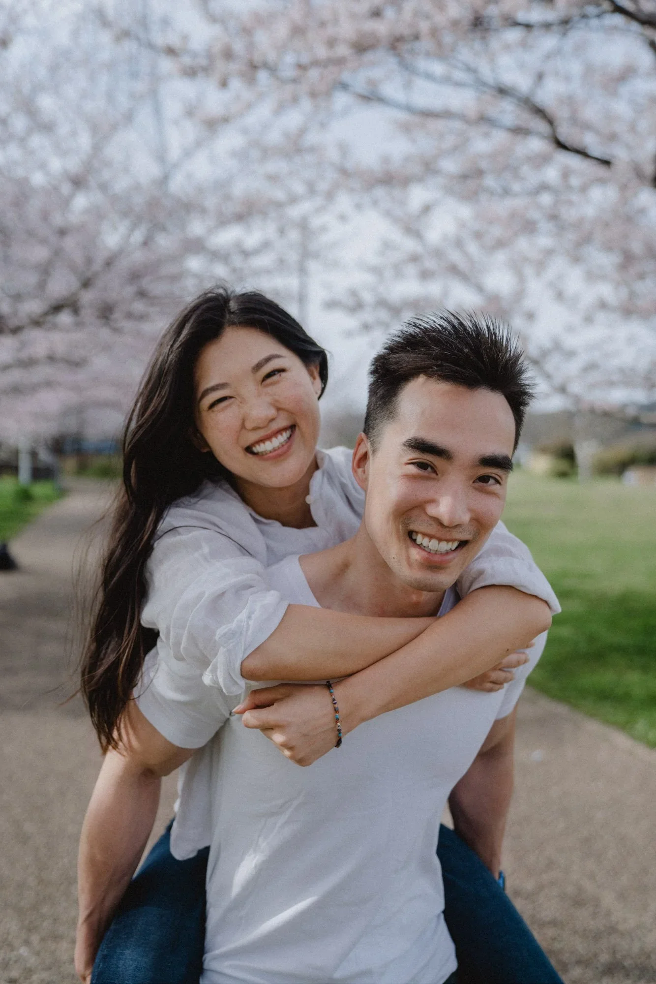 A smiling young woman and man enjoying a piggyback ride outdoors during cherry blossom season.