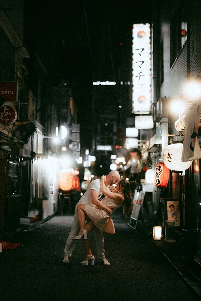 A couple dances intimately in a narrow, brightly lit alley at night, surrounded by Japanese lanterns and signs.
