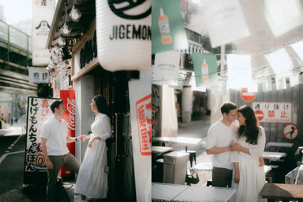 A young man and woman in white clothing share a moment and smile at each other on a street with Japanese signage, banners, and hanging paper lanterns.