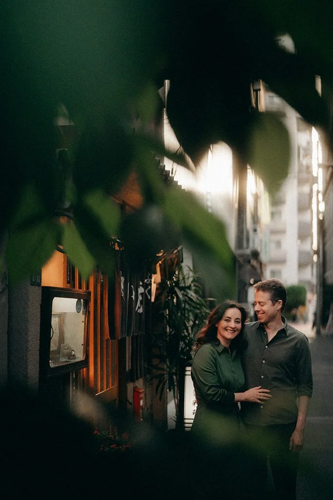 A smiling woman and man walking on a city street at sunset, wrapped in a close embrace, with buildings and trees in the background.