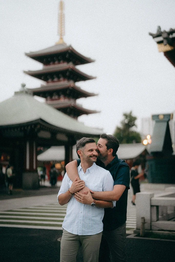 Two men hugging and smiling in front of a traditional Japanese pagoda at dusk.
