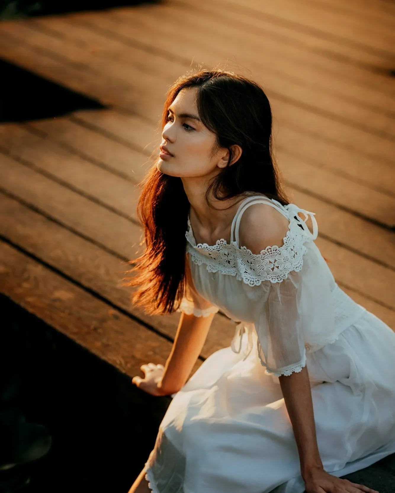A woman with long dark hair sitting on a wooden deck during sunset, wearing a white dress with lace details on the shoulders, gazing thoughtfully into the distance.